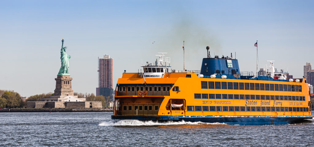 Get to Delightful Destinations in Staten Island via the Staten Island Ferry. Seen: Yellow Staten Island Ferry passing Statue of Liberty in New York Harbor.