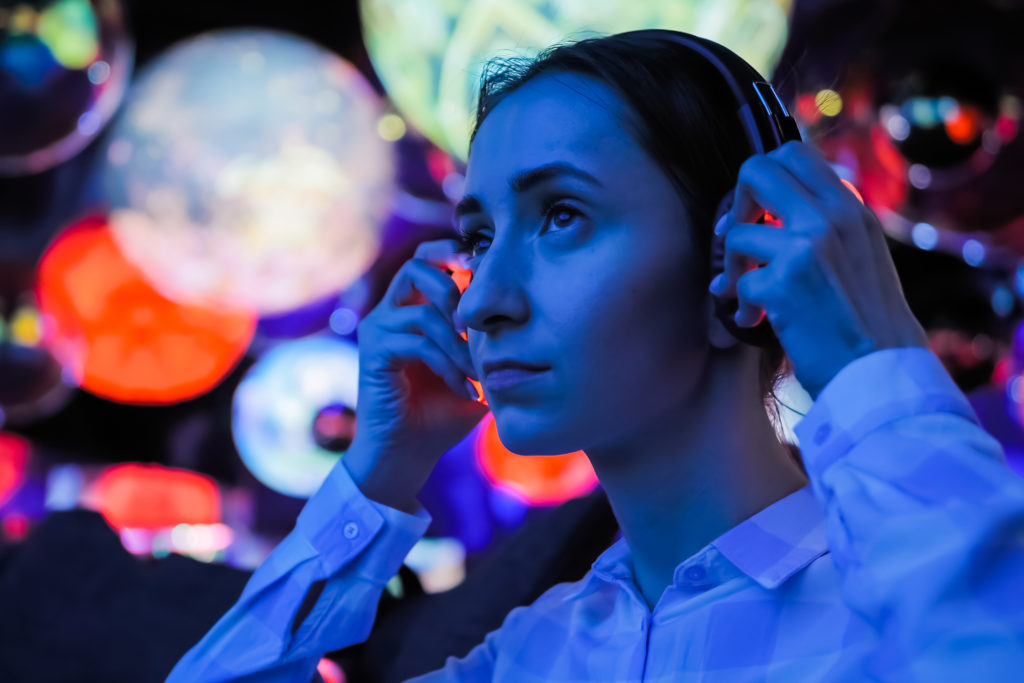 Woman wearing wireless black headphones and looking around in dark room of interactive exhibition of museum with colorful illumination