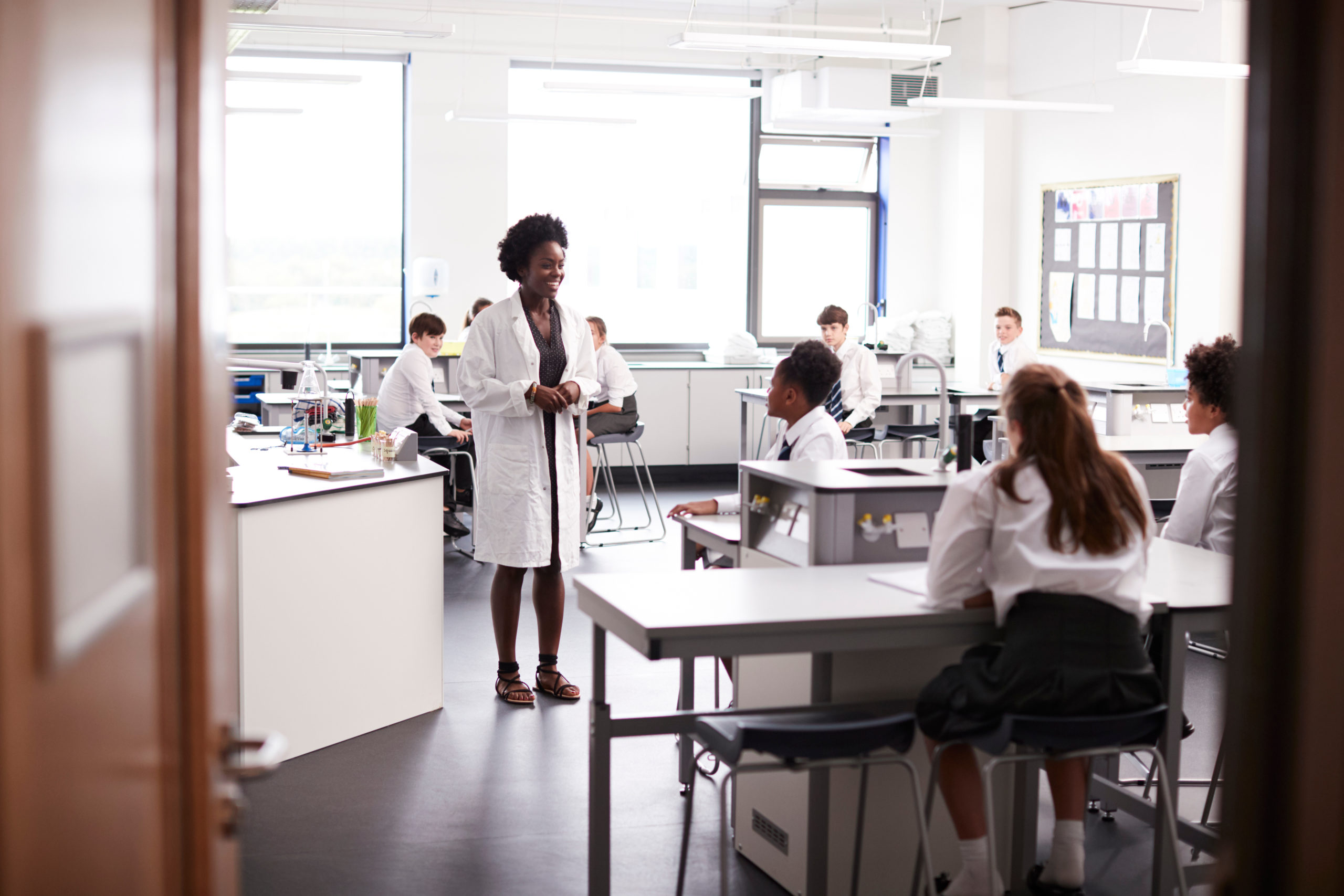 Teacher in a High School Science Lab Class, wearing a lab coat, teaching students who are wearing uniforms of white shirts and dark skirts and pants